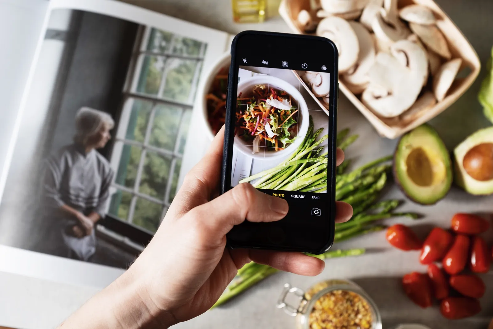 A hand holds a phone, photographing a salad on a sunny kitchen counter beside an open book